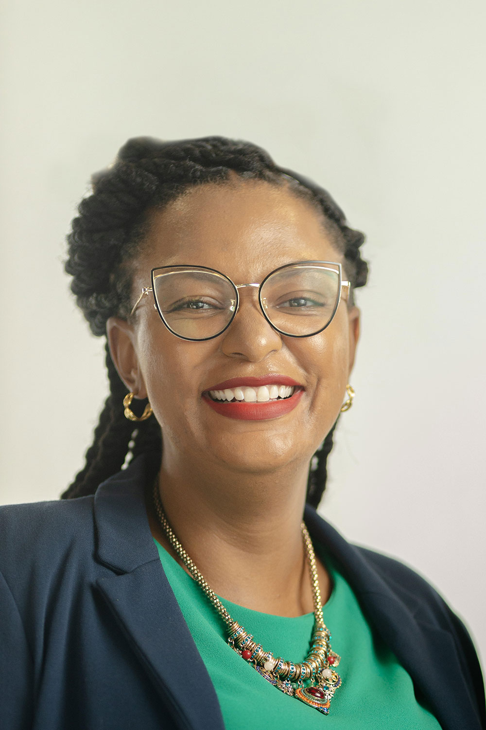 Smiling woman with braided hair, glasses, gold hoop earrings, and colorful necklace in blazer and green top.